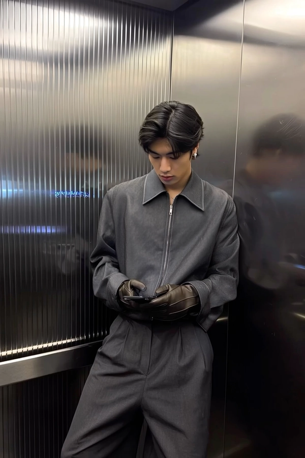 Cool graphite hues of a zipped mockneck shirt shift against the gentle flare of deep obsidian pants, the subtle grain of supple leather gloves catching snapped light as he leans with relaxed poise against a mirrored elevator wall detailed by vertical ridges. The flash picks up each groove like a pixelated glitch, fragmenting reflections in static ripples that dance quietly around his sharp jawline and neatly kept hair. The phone, held low at waist height with a faint smudge on the lens, captures the slight slope of his shoulder and the unbothered tilt of his head as he gazes down at the screen, fingers poised near the sleek control panel. Overhead fluorescents cast steady clarity, mixing with a digital shimmer that brings out textures—a weave of knit on the mockneck, the satiny tired shine of fabric catching the elevator’s cool steel vibe. There is an effortless coolness in his stance, folding simplicity and subtle details into a moment caught between floors—a silent nod to casual refinement and urban edge.
—elevator mirror capture, urban chic on iPhone