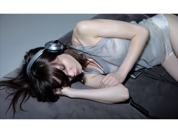A pale, ethereal glow fractures across translucent skin, a whisper of cold key light splintering over the curve of a sleeveless, faded cotton tank top—a whisper of lived-in softness. The model reclines languidly on a matte charcoal velvet couch, arm draped like a veil over her eyes, her tousled hair spilling into obscurity. Her expression is one of quiet surrender, a hushed resistance to waking, while vintage over-ear headphones with cracked leather offer a tangible relic of analog intimacy.

A scatter of tangled black cables and the muted silver edge of a laptop hover just out of focus, their insignificance enhancing the arresting vulnerability of the figure. The harsh digital flash slices unevenly, casting spectral shadows and imbuing skin with an unsettling translucence, as if caught between dream and wakefulness. This interplay of stark illumination and shadow clamps reality in a fleeting pause, breath caught beneath the gentle weight of insomnia.

The tone is cool, washed in a muted palette of ghostly whites and shadowed grays, punctuated by the warm grain of worn fabric threads and the textured fragility of weathered leather. The frame is tightly cropped from above, shot on a 85mm prime at mid-aperture, compressing the space to sculpt a fragile stillness—each wrinkle, each pore magnified to intimate clarity.

The entire composition channels a low-key, near-noir sensibility with a hint of surreal flash tableau, a haunting echo of vintage film stocks rendered through sharp digital precision. The result is a resolute arrest of ephemeral stillness—high-fashion editorial, hyper-real texture fidelity