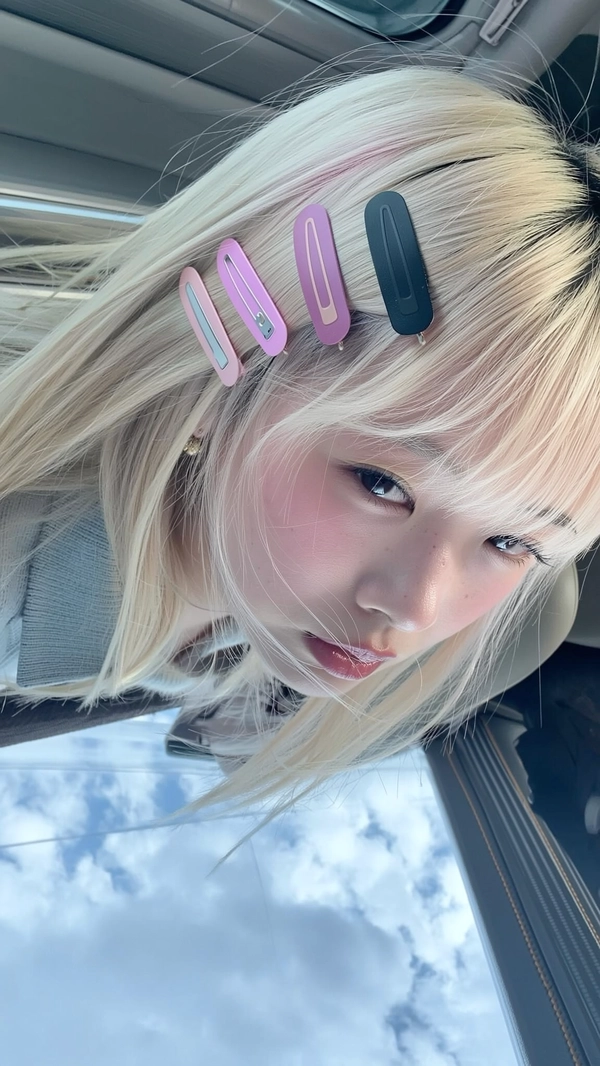 Pastel pink and matte black clips scatter symmetrically across platinum blonde bangs, their soft pastel tones catching a cool, overcast street light that filters through scattered clouds. The pale girl’s calm stare peers into the frame, eyes reflecting wistful nostalgia with the faintest dewy glow on her cheeks. Fly-away strands gently skim her porcelain skin, the smooth texture disrupted only by subtle skin pores and wisps that frame her delicate face. The faint grain of a wool sweater collar brushes her neck, a seatbelt edge visible at the frame’s corner grounding the moment in quiet city transit. The iPhone's slight high-angle tilt reveals a dreamy softness interspersed with crisp clips that steal the spotlight—authentic iPhone close-up, statement hair-clip focus