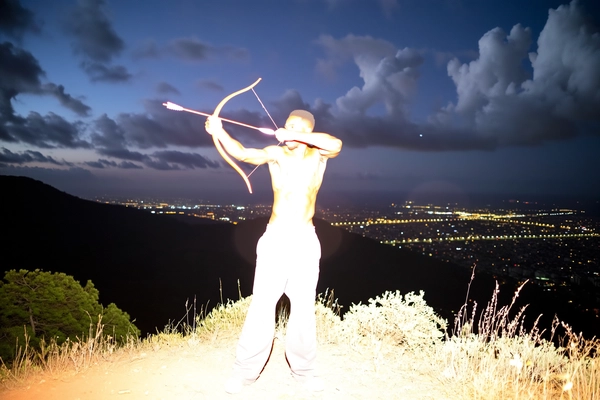 A spontaneous nighttime iPhone flash snapshot of an Afro-Latin man standing shirtless on a hilltop viewpoint. He wears loose linen pants and aims a toy bow and arrow into the air. The intense, direct flash overexposes and bleaches out his muscular body into a glowing white silhouette, obscuring facial and clothing details completely. Dramatic clouds and distant city lights form a dark, textured background. The framing is casual and imperfect, emphasizing the surreal and spontaneous nature of the moment.