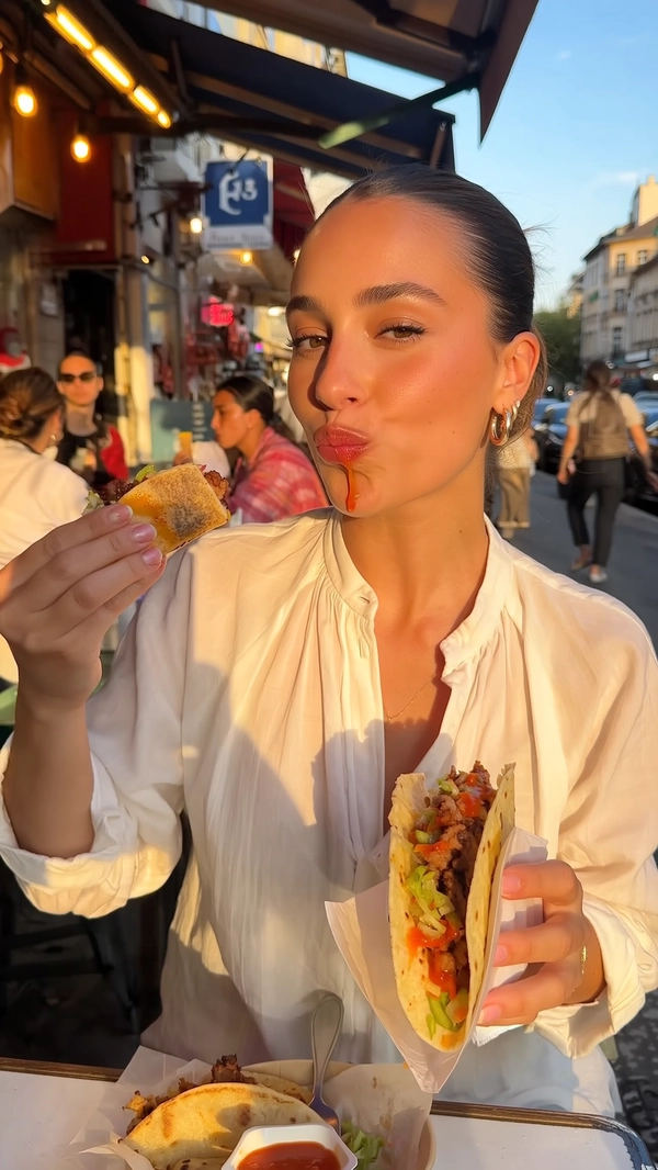 Bathed in the golden hour’s honeyed light, a striking young woman with sleek chestnut hair tucked behind one ear sits calmly at a bustling street food stand. Her attire is effortlessly chic: a soft, cream-colored blouse coupled with delicate gold hoops catching the light. The gentle warmth of the setting sun filters through nearby trees, casting dappled shadows and an amber glow over her flawless skin, which subtly reflects the soft sheen of a barely-there lip gloss. Her serene, half-lidded eyes meet the camera with a neutral poise, the calmness underlining a quiet elegance.

In her hand, a generously filled taco is lifted casually toward her parted lips, a careful yet unhurried bite about to be taken. The layers of charred meat, crunchy lettuce, and vibrant salsa peek from the slightly folded tortilla, the bright red of the sauce glistening with fresh juiciness. A tiny dribble of salsa clings delicately to the corner of her mouth, a tactile reminder of the taco's messy richness, subtly breaking the composed stillness.

The scene stretches beyond her — a softly blurred background of an urban street corner and fellow patrons, hinting at the lively but intimate afternoon street food atmosphere. The gentle hum of the city mingles invisibly with the warm air, the soft textures of woven fabric and fresh food scents almost tangible. The angle is a near-knee level, casually capturing her from an arm's length perspective, lending an authentic iPhone snapshot feel.

Her posture and expression hold steady, embodying a fashion-forward calm amid the vibrant chaos of street dining. The interplay of warm light, glossy salsa, and tactile food textures create a refined yet spontaneous moment of quiet indulgence, perfectly resembling a candid iPhone photo.