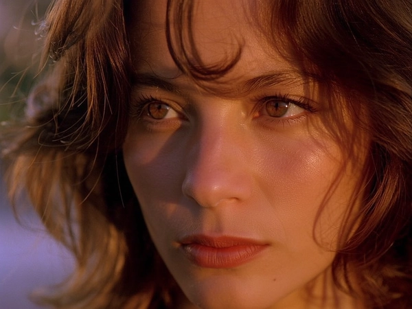 Bathed in the soft, warm glow of late afternoon light, a close-up of a woman's face reveals delicate details—her porcelain skin glows with a gentle sheen, framed by loose, tousled waves of chestnut hair catching the light like silk. Her eyes, wide and contemplative, shimmer with quiet introspection, while a single stray lock brushes softly against her cheek, suggesting a moment suspended between thought and feeling. The muted amber hues cascade gently across her features, lending warmth to the natural texture of her clear skin and the faint blush of her lips. A soft background blur cradles her profile in intimate focus, evoking a cinematic tenderness that invites the viewer into her private world. Fine grains settle subtly across the frame, enriching the tactile realism with a whisper of nostalgic depth, as if captured on delicate vintage film—evoking both presence and poetry in a singular, unforgettable gaze.