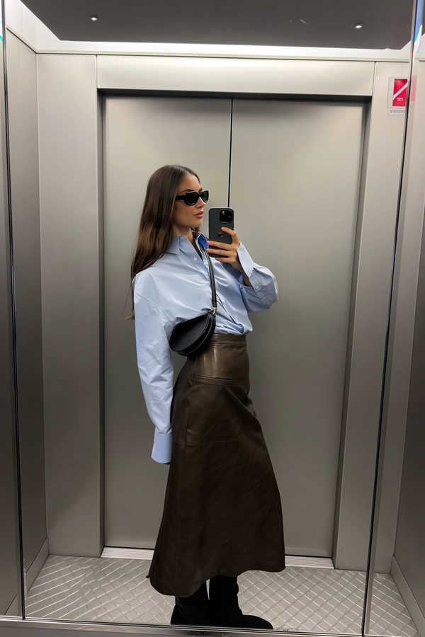 Cool grey panels rise behind her, the silver checkered floor anchoring the frame where she leans just so, in her oversized crisp shirt paired with a leather midi skirt that catches soft wrinkles and the subtle satin sheen of touch-worn leather. Her circular bag swings gently by her side as reflection finds her black sunglasses twice—once caught in the mirror's honest glass, again as a ghost glinting from the brushed steel panel. The phone is held waist-high, fingers poised near a faint smudge on the glass; overhead LEDs hum a cool, even glow that smooths skin texture with a whisper of noise. The composition crops her crown out, and the pause in the closing doors presses a calm motion into the moment—elevator mirror capture, urban chic on iPhone