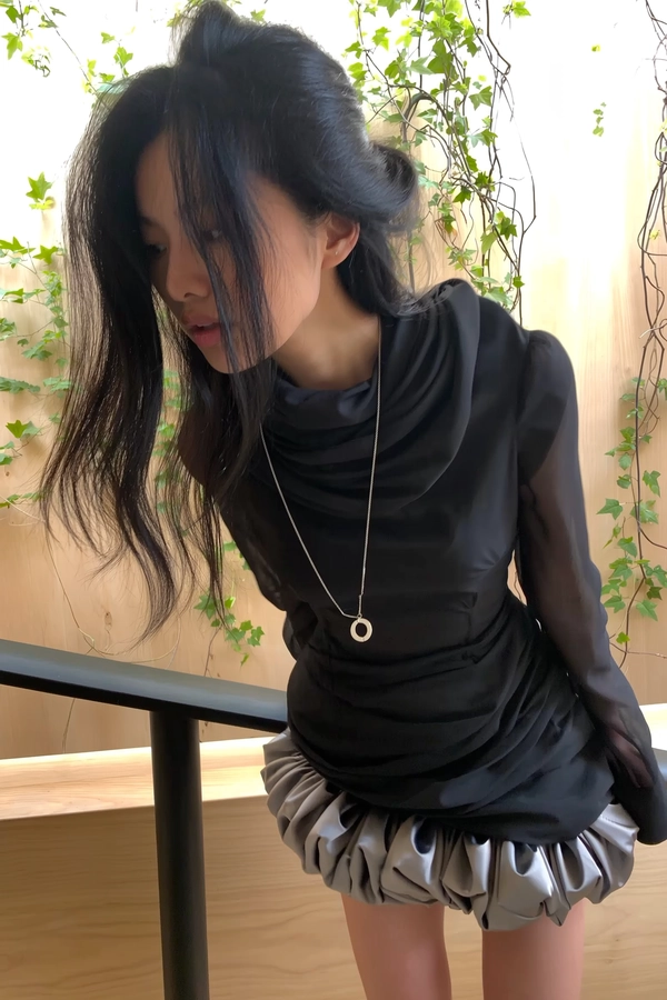 A casually candid indoor portrait spontaneously captured on iPhone showing a woman with thick, heavy black hair styled naturally with loose strands softly framing her face. She wears a sculptural black blouse featuring exaggerated neck wrapping, layered minimalistic textures, and a simple circular pendant necklace conveying refined Japandi elegance. Her bottom half is partially obscured by a playful yet tense ballooned satin miniskirt adding subtle contrast in texture and shape. The softly diffused indoor daylight filters gently across warm, minimalistic wooden surfaces and a background of delicate leaves and vines intertwined with a simple black railing, emphasizing tactile textures and natural skin details. The framing is casually tilted and intimately close, capturing an unposed, spontaneous moment with subtle shadowing and ambient reflections enhancing authentic iPhone candidness.