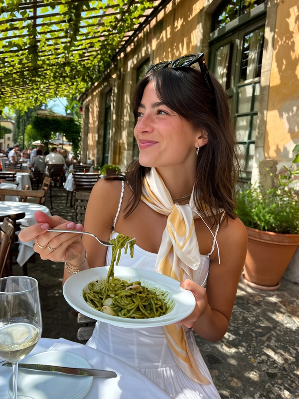 A candid photo of a young woman sitting on a shaded terrace at an authentic Italian trattoria, casually enjoying a plate of pesto pasta. She wears a chic white corset dress paired with a soft pastel silk scarf loosely tied around her neck and vintage-style oversized sunglasses resting on her head. Her dark hair is softly tousled, and her relaxed smile reflects genuine pleasure. The terrace features rustic wooden tables and chairs with weathered stone walls adorned with creeping green vines and terracotta pots. Soft natural daylight filters through leafy overhead shade, casting gentle shadows that warm her sun-kissed skin and accentuate the textured fabric of her dress and silk scarf. The pesto pasta’s vibrant green contrasts with the earthy tones around her. The image is framed with a slight natural tilt and casual cropping that captures the spontaneous intimacy and tactile realism characteristic of an iPhone candid snapshot, embodying the chic dolce vita atmosphere of an Italian summer afternoon.