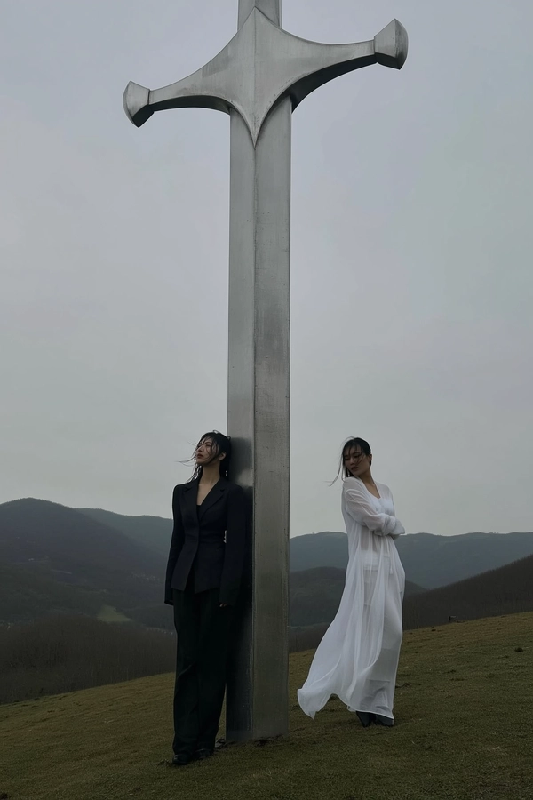 A spontaneously captured, muted-toned iPhone photograph depicting two women standing solemnly on a softly sloping hillside dominated by a colossal, vertically embedded metallic sword. One woman wears a structured black outfit with subtle gothic undertones, her hair casually tousled and slightly damp, revealing genuine skin texture with minimal makeup. The other woman is dressed in a flowing white garment that moves gently in the cold breeze, her semi-transparent fabric catching the diffused light under an overcast grey sky. Both exhibit introspective, serene expressions, unposed and quietly reflective. The distant mountain range under a soft, fog-filtered daylight enhances the surreal atmosphere. The massive sword’s worn metallic surface softly reflects the muted landscape hues. The composition is off-center, slightly tilted, capturing scale contrast between the monumental blade and the human figures, evoking a cinematic, melancholic mood characteristic of authentic spontaneous iPhone photography.