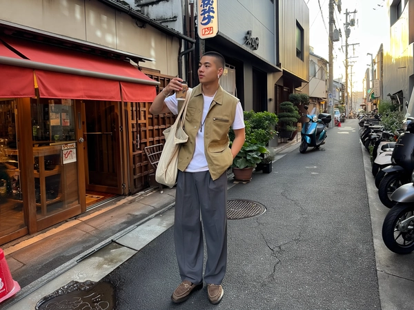 Sunlit soft afternoon hues wrap a young man lingering near a nimble soba shop’s back alley, where a red noren curtain sways, teasing the warm breeze. He drifts casually in a sand-hued utility vest layered over a crisp white tee, wide slate trousers folding over scuffed leather derbies. His buzzed haircut gleams faintly beneath a subtle silver earring glint, while a rugged canvas tote hangs loose from one shoulder. Half-lifting a chilled Boss Coffee can, he blends into Tokyo’s layered quiet chaos: scooters parked unevenly, potted greens juxtaposed against overhead snarls of wires. The cracked alley’s asphalt bears faint puddle gleams, catching bits of fading sunlight that the iPhone lens renders with gentle shadows and an amber warmth. Framed from a slightly tilted hip-level angle, edges crop unpredictably as if snapped on impulse, while background scooters blur softly, conveying casual urban texture. The moment hangs suspended, effortlessly real and inviting, alive in Tokyo’s intimate pulse—Tokyo street snap, shot on iPhone