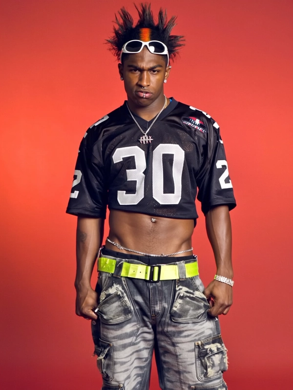 He’s standing against a saturated red studio backdrop, posture relaxed with one shoulder slightly dropped and his arms resting at his sides like he’s not trying to hold anything. He’s wearing a cropped black football jersey with oversized white numbering, the mesh fabric revealing a sliver of his toned torso and a silver belly chain hugging tight across his skin. His pants are low-rise cargo denim in icy gray with bleach stains and exaggerated pockets, cinched with a neon yellow industrial belt that drapes halfway to his knee. His hair is dyed jet black with a fire-orange streak running through the front, styled into sharp upward spikes. A pair of white wraparound sunglasses rest on his forehead, and his lip is pierced with a small hoop that glints under the warm artificial lighting. He stares forward with a dead-serious expression, equal parts aggressive and bored, like he’s stepped out of a futuristic sports team promo with too much attitude to care.
—casual candid early-2000s Y2K snapshot, grainy softness
