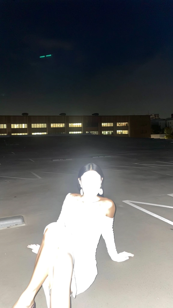 A spontaneous nighttime snapshot on an empty rooftop parking lot, captured with intense direct flash from an iPhone. A Southeast Asian woman lounges with legs crossed, wearing an off-shoulder dress and large earrings, but her skin and features are completely bleached out and glowing white from the flash, leaving only ghostly outlines visible. The distant office windows twinkle faintly behind her in the dark. The framing is casual and imperfect, emphasizing the surreal, candid moment and ethereal figure created by the overexposure.