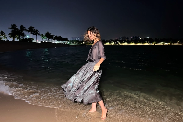 Under a sudden iPhone flash, she stands just where the surf and sand meet, the midi-skirt and fluid blouse catching the gentle seaside breeze, woven folds reflecting faint glimmers of distant city lights and moon's shy glow. Her bare feet nestle softly in the warm, textured sand, illuminated subtly by distant streetlamps that drift behind her, palms and skyscrapers whispering quiet stories to the night. Damp tendrils of wavy hair move gently, framing a serene, turned face softened by reflective air, while the expansive darkness spills across two-thirds of the frame in velvety repose. A delicate blur trails her skirt's hem, contrasting with the crisp focus of her poised stance, embodying timeless grace and effortless charm—flash-lit elegant beach snapshot, captured on iPhone.