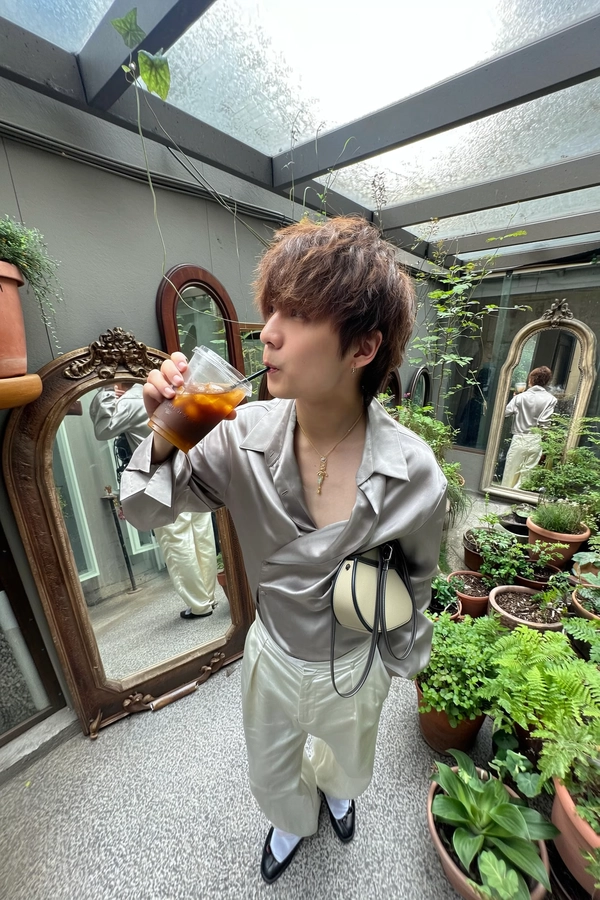An elevated wide-angle photo realistically taken on an iPhone features a tall, androgynous figure casually sipping an iced beverage while standing in a whimsically artistic courtyard densely arranged with antique, ornately framed mirrors and varied lush potted greenery. They look directly upward into the camera with a calm, natural neutral expression, their hair styled in a subtly tousled, gender-neutral cut. Their uniquely reinterpreted outfit includes an asymmetric, smooth-textured satin blouse in a soft metallic hue with creative sleeve and collar detailing, tucked into high-waisted silk trousers featuring a fluid drape and gentle sheen distinctively different from described examples. They carry an imaginatively designed baguette-shaped purse in complementary tones, accented with unconventional hardware and modern minimalist straps. The surrounding antique mirrors craft layered, softly distorted reflections contributing to an ethereal atmosphere under soft natural daylight filtering gently through. Visible textures encompass light fabric creases, realistic silk sheen, delicate hair strands, slightly dewy skin surfaces, and richly detailed plant leaves with subtle imperfections. The composition embraces authentic exaggerated wide-angle distortion and casual elevated camera angle typical of iPhone snapshots, capturing the subject’s engaging upward gaze and natural posture within a lively, textured courtyard scene. This spontaneous snapshot embodies a stylishly playful, genuinely neutral moment enhanced by the dreamy mirror reflections and vibrant urban greenery, perfectly reflecting the dynamic and authentic aesthetic of wide-angle iPhone photography.