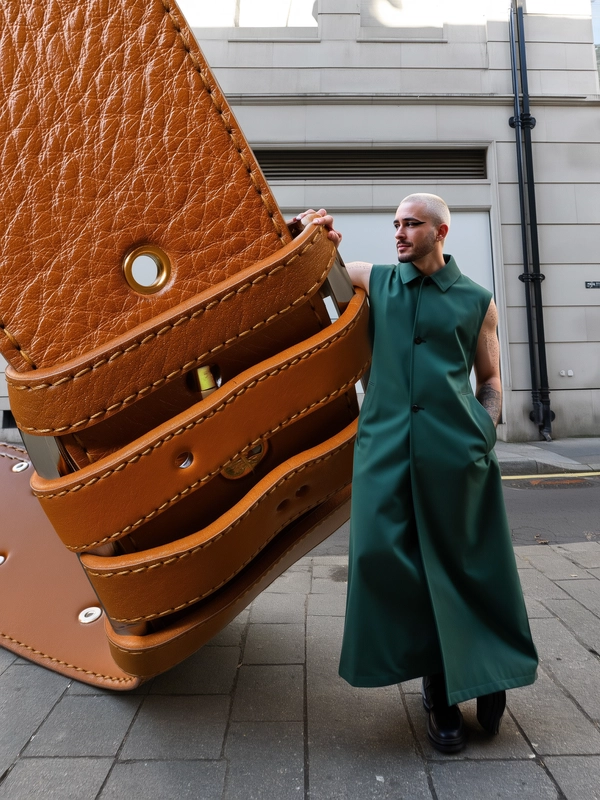 A spontaneous, candid iPhone-style photo of a man leaning casually against an enormous caramel leather belt buckle that curls dramatically through the urban scene, resembling architectural curves. The man sports an icy-white buzzcut, sharply defined geometric black eyeliner, and a sleek floor-length sleeveless trench coat in deep forest green that mirrors the belt’s flowing lines. The oversized belt features finely detailed leather grains, subtle stitching, and glossy highlights realistic to high-quality caramel leather. He stands on a lightly cracked concrete sidewalk outside a modern building facade with subtle urban graffiti and metal fixtures. Gentle natural daylight softly illuminates the textures of his coat’s fabric weave, the belt's leather surface, and his skin with visible pore details. The composition has a slight tilt and off-center framing typical of spontaneous iPhone snapshots, casually integrating this surreal oversized architectural belt element in a believable candid street photograph style.