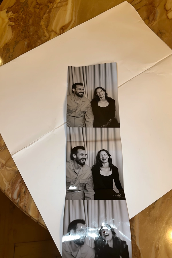 A vertical black-and-white photostrip lays casually over folded parchment paper atop a warm marble table, slight bends tracing its length. The muted curtain backdrop in soft beige fabric folds gently behind two figures: a man in a minimal tan jumpsuit and a woman clad in a black fitted top, leather gloves adding texture. Frames unfold naturally—the pair averted gazes in the first, the man’s smirk rivaling the woman's tilted head in the next, culminating in both mid-laugh, radiating spontaneous joy. Subtle iPhone flash reflections and gentle shadows ripple softly, surface grain and faint fingerprint smudges tracing the strip's vintage authenticity. An overhead, gently skewed view echoes the intimacy of a candid capture—contemporary photobooth snapshot, shot on iPhone.
