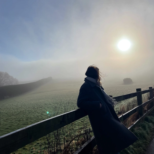 A lone figure wrapped in a thick wool coat and knitted scarf leans gently against a weathered wooden fence, shoulders relaxed yet inward, amidst a fog-wrapped countryside where rolling meadows fade softly into mist. The damp wood glistens faintly with moisture, and his exhaled breath rises as pale whispers in the cool dawn air. The mist diffuses the muted greens and tawny earth beyond, engulfing distant hedgerows in grey softness. Strands of hair peek from beneath the scarf, caught in a subtle breeze that nudges the coat hem. Grain lifts in the shadowed fence boards, while a pale sun bursts weakly behind the clouded veil, casting a gentle halo that the iPhone lens catches with informal tilt from waist height. Wide negative space swells around him, letting quiet linger, suspended between earth and mist—mistbound reverie, shot on iPhone