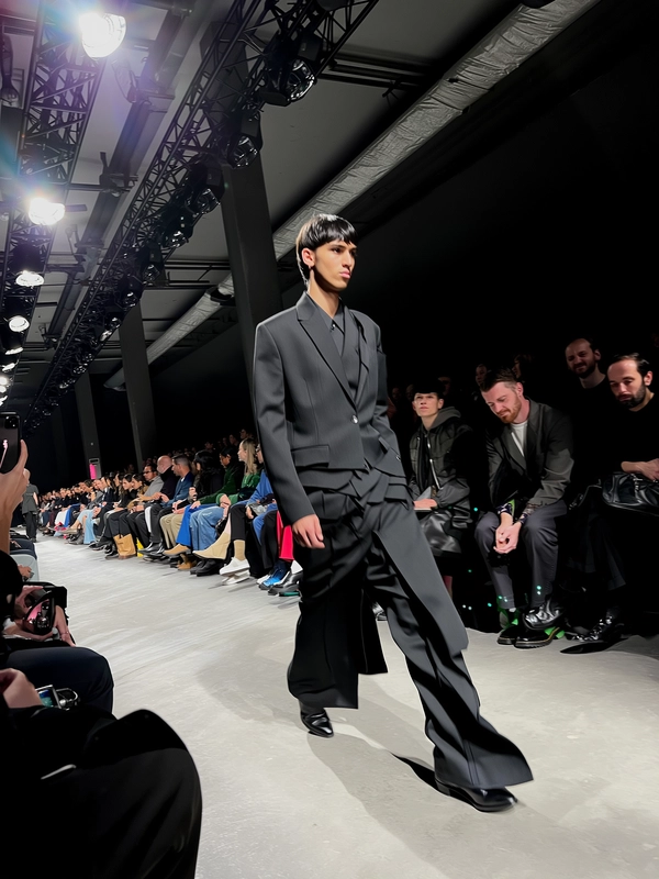 A male model wrapped in layered, all-black tailoring strides confidently down the narrow warehouse runway, the structured garments’ matte and subtle textures absorbing the harsh top spotlight that casts sharp shadows and ghostly highlights over his angular frame. The dim venue reveals a sparse layout with a single row of minimalist chairs flanking the concrete runway, occupied by focused spectators whose faces flicker in the low ambient glow. Overhead, industrial trusses and exposed ducts cut a raw silhouette against the background, while the intense spotlight bleeds mildly at the edges, giving the model an almost ethereal glow. Slight motion blur trails his brisk steps, contrasting with crisp details in the refined fabric folds and the faint leather glint on pointed boots. The iPhone’s sensor captures rough grain in deep shadows and faint bloom over the light, as a cropped, slightly tilted angle from the front row emphasizes the intimacy and immediacy of the moment—contemporary runway snapshot, shot on iPhone