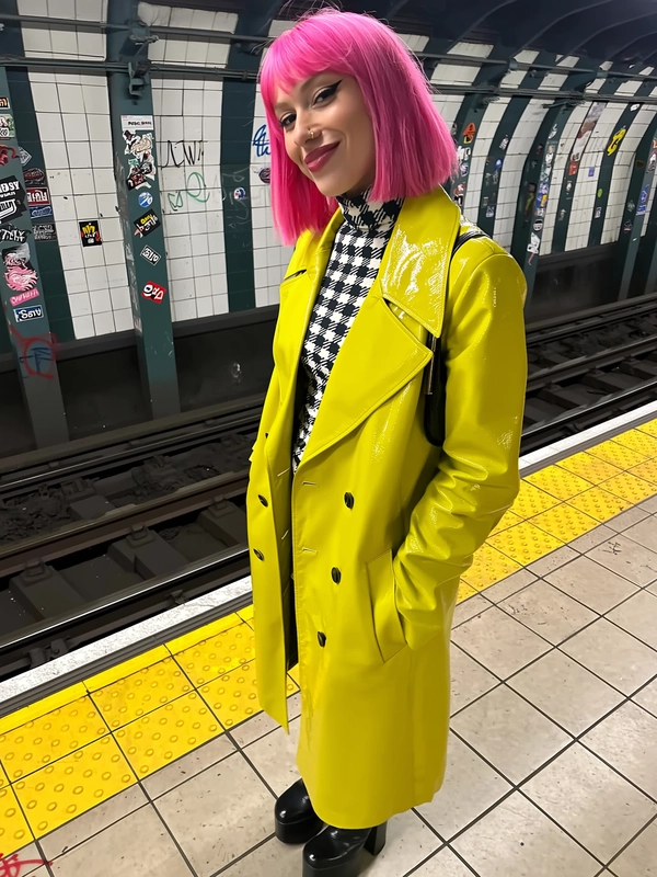 A spontaneous candid photo of a striking woman with a candy-pink bob hairstyle and sharp eyeliner, standing casually on a tiled subway platform covered with sticker graffiti. She wears a vivid yellow patent-leather trench coat over a black-and-white checker-print turtleneck, paired with platform boots, all rendered with natural subway lighting that highlights the glossy texture of the trench and the fabric grain of her top. The gritty background tiles and graffiti add raw urban texture with soft, realistic shadows typical of an iPhone capture. The composition is casually framed, with a slightly tilted angle emphasizing genuine, unposed authenticity and relaxed confidence in an everyday city environment.