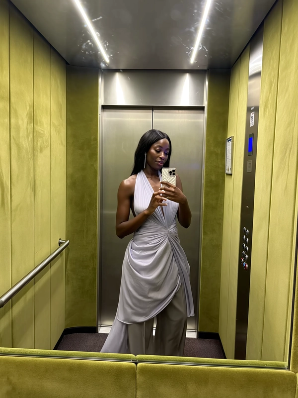 A delicate spread of moss green suede wraps the elevator cabin, its calming embrace softening the glow of the warm overhead lights. She stands centered, a long halter-neck dress in whispered chiffon layered over draping wide suit trousers that sway with an effortless rhythm. The phone is cradled gently at chest height, her fingers arranged with the quiet precision of a gallery catalog, as if each movement is a curated exhibition of relaxed refinement. A pale sheen catches the folds of fabric; her skin texture hums under the soft light, and a faint fingerprint smudge lingers just below the lens. The frame cuts slightly above the waist, as the elevator hums softly—a private moment of poised ease between floors as the closing doors hint at movement beyond—elevator mirror capture, urban chic on iPhone