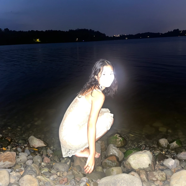 A nighttime snapshot taken with an iPhone flash at the shore of a dark lake. A young East Asian woman crouches on jagged stones, wearing a vintage lace slip dress. The intense, direct flash completely bleaches her facial features and details of her dress, rendering her a ghostly white silhouette against the inky black water. The spontaneous framing and natural wet hair emphasize the candid, surreal moment.