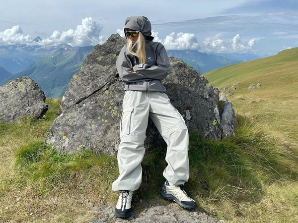A candid, full-body image spontaneously captured with an iPhone, portraying a person dressed in a functional, outdoor-brand Arc'teryx's gear reflecting the gorpcore aesthetic. The individual wears a silver gray hooded technical jacket with structured seams, multiple zippers, and chest pockets, the durable, weather-resistant fabric realistically showing creases and slight signs of use. Their light blonde hair flows naturally from under a hood pulled up casually. They sport oversized off-white cargo pants with wide legs and articulated panels, creating a sculptural silhouette, paired with off-white and black trail shoes featuring thick soles and rugged detailing. The person stands relaxed with arms crossed against a textured rocky surface on a grassy alpine meadow beneath a cloudy mountain ridge. Softly diffused daylight with partial clouds enhances the fabric textures, rock details, and surrounding wild grass, while reflective lenses on wraparound sunglasses capture subtle mountain reflections. The framing is casually tilted, invoking the genuine spontaneity and intimate style typical of trendy iPhone outdoor photography.