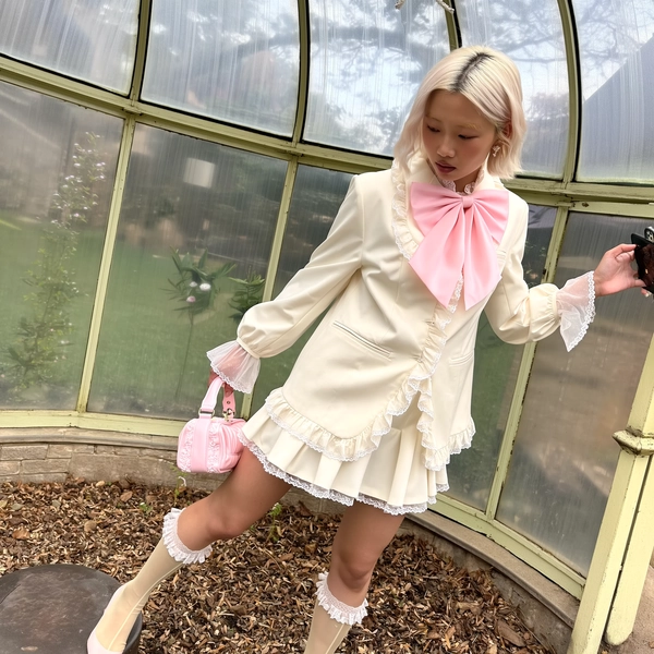 A gently candid moment captured on an iPhone, featuring a young woman with softly styled platinum hair, dressed in a whimsical Jenny Fax tailored mini dress ensemble in creamy white, adorned with soft ruffles along the hem and subtle scalloped lace trims at the cuffs and neckline. She wears sheer puff sleeves layered beneath the structured blazer, complemented by delicate pastel pink accents including a dainty handbag and a voluminous oversized bow at the collar. Her look is completed with sheer lace-trimmed socks peeking above translucent heeled boots. Set within an intimate, softly aged glass conservatory filled with diffused daylight that bathes the airy fabric textures and vintage-inspired architectural details in gentle warmth. The tender lace and delicate ruffle textures shimmer subtly against the softly reflective glass surfaces. Framed from a slightly elevated and tilted angle capturing the model’s quietly introspective expression and graceful poise, the composition balances casual charm with refined femininity, evoking a dreamlike sense of whimsical vintage elegance.