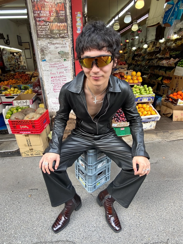 A spontaneously authentic wide-angle iPhone-style photograph showcases a fashionable man casually seated on a stack of plastic crates positioned just outside a bustling urban market. He looks directly up into the camera with a relaxed neutral expression, his hairstyle neatly tousled yet natural. His outfit creatively reimagines a sleek leather one-piece with a partially unzipped front, distinguished by original design elements such as asymmetrical seams and layered texture, paired with distinctive crocodile-pattern ankle boots featuring artistic color gradations and sculptural soles—far from typical or duplicated styles. He accessorizes with large tinted visor-style sunglasses unique in reflective gold hues, enhancing his bold yet refined appearance. Behind him, the lively street scene brims with intricately detailed cardboard boxes bearing faded textures, vibrantly fresh fruit stands rich in natural color variety, and weathered wall posters layered with hand-painted notes and scribbles that add urban authenticity. The daylight softly diffuses over the scene, creating natural shadows and subtle reflections on surfaces. The composition utilizes a pronounced elevated camera angle, wide perspective, and casual framing characteristic of casual iPhone shots, notably exaggerating upper body proportions while compressing the lower body, producing a dynamic, playful yet natural wide-angle distortion. Textural authenticity emphasizes realistic skin pores, individual hair strands gently catching light, soft creases in leather fabric, worn-in boot textures, and gritty urban surfaces, all harmonizing to evoke an engaging, spontaneous moment of urban fashion and activity captured with genuine iPhone photographic charm.