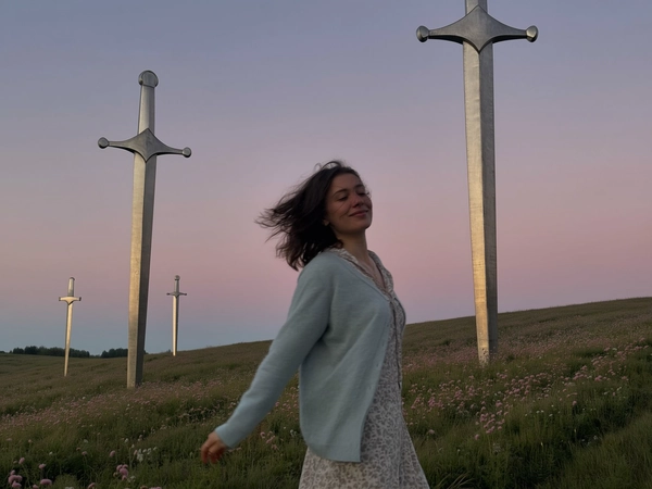 A spontaneously captured, muted-toned and quietly joyful iPhone photograph of a Lithuanian woman spinning gently in a wildflower field at dusk. She wears a soft pastel cardigan layered over a semi-transparent patterned blouse, her hair lightly tousled by the gentle evening breeze, exhibiting subtle wind-tousled texture and natural minimal makeup with visible authentic skin details. The sky behind her fades in muted pink hues under a soft twilight diffusion, blending into an expansive landscape of softly sloping grassy hills punctuated by immense, vertically embedded monumental metal sword sculptures. The swords’ worn metallic surfaces gently reflect the ambient dimming light filtered through delicate haze. The overall color palette is subdued with dusty blues, faded greens, and pale pinks, evoking a cinematic, surreal atmosphere. The composition feels intuitively off-center and casually tilted, capturing the natural scale contrast between her spontaneous movement and the towering surreal swords, emphasizing quiet introspection and emotional storytelling through this atmospheric scenery.