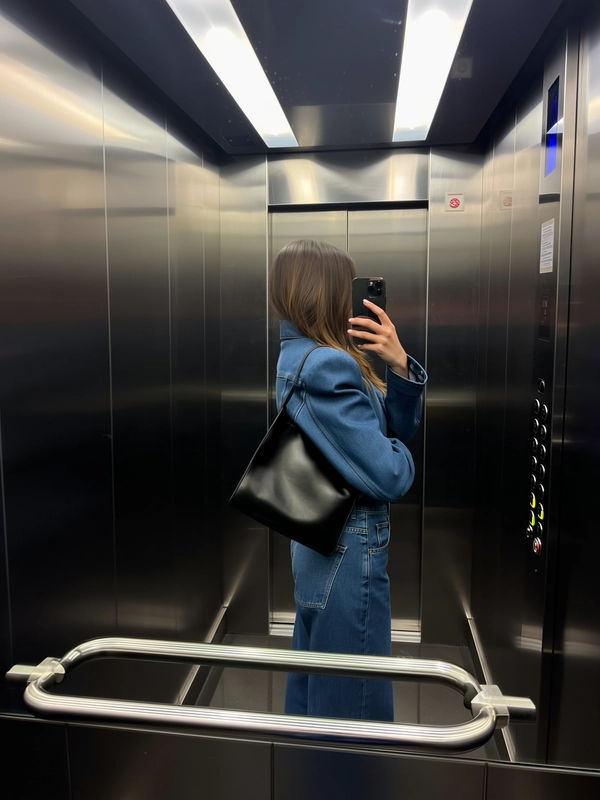 The cool blue of structured denim sharpens her silhouette even in the tight confines of a mirrorless, chrome-paneled elevator where her stiff leather clutch presses casually against her hip. An exaggerated shoulder line lends architectural resolve to the softly fluid knit beneath, while her face appears only in a fleeting, distorted glimpse within a chrome control panel’s reflection. Fingers hover near a button as the phone tilts chest-high, catching the satin sheen of her clutch and the subtle fingerprint trails on the polished metal. The overhead fluorescents flicker gently, lending a muted glow that softens the rigid space, and a slight barrel reflection curves the cramped frame of this private ascent. Unbothered and quietly confident, she leans into the small moment, the city's pulse faint behind the sealed doors. —elevator mirror capture, urban chic on iPhone