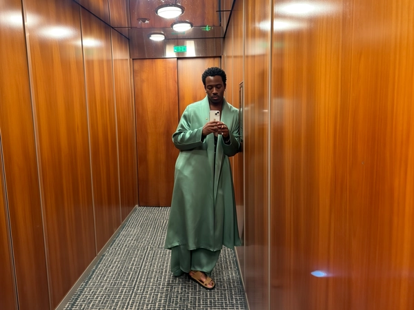 Soft muted green separates in fluid, oversized draping weave gently catch the warm gleam of wood-paneled walls, while a mirrored ceiling above fractures the faint hop of evening light. His sandals rest grounded on the patterned floor, toes barely clipped by the frame, as his eyes glide just past the iPhone held steady at chest height. The smooth satin sheen of his attire contrasts with the rich wood’s grain, occasional fingerprint streaks blurring the glass around the sensor. His relaxed jaw and barely-there crease between brows read unbothered, almost detached, as if caught mid-thought while buttons near his fingertip hum in wait. The room breathes a quiet depth through faint reflections scattered in the mirrored ceiling’s soft edges, wrapping an offhand elegance into this suspended moment—elevator mirror capture, urban chic on iPhone