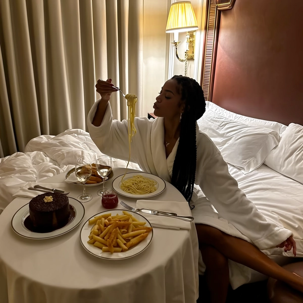 Draped loosely in a plush white bathrobe, her long braids cascade down one shoulder as she perches at the edge of a lush, unmade bed. The warm glow of a vintage wall sconce spills softly over creamy pasta strands lifted mid-twirl from a delicate fork, their sheen catching glimmers against the fine porcelain plates arrayed before her. A round table dressed in crisp white linens holds twin plates of glossy chocolate cake with rich sauce pools, golden fries, and tiny jars of jam—traces of a slow breakfast folded quietly into this rich evening tableau. Thick drapery muffles the room into a private hush, where ambient light pools generously yet shadows cling warmly along the bedspread’s folds. The iPhone's gentle sensor grain wraps this quiet, elegant pause in a tactile luxury, framing the relaxed moment like a whisper of an indulgent slow night in a high-thread-count haven. —leisure-suite snapshot, shot on iPhone