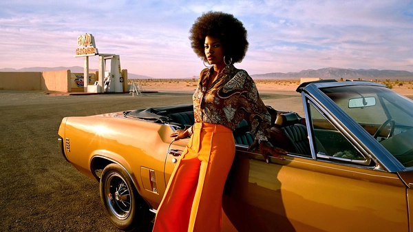 In the golden haze of late afternoon, a confident Black woman with a voluminous afro leans nonchalantly against a mustard-yellow convertible from the 1970s, the car’s glossy finish catching the sun’s warming light. Her high-waisted orange flared trousers flare wide and bold, contrasting effortlessly with the muted earth tones of her intricately patterned paisley shirt, tucked neatly to highlight her slender waist. Oversized gold hoop earrings swing lightly as the desert breeze stirs the curls framing her serene yet commanding face, full lips pressed into an expression of unshakable poise.

The setting—a retro-style gas station with classic pumps and faded signage—sits under a vast sky streaked with pastel Fuji dusk hues that bleed softly across the horizon, bathing the scene in nostalgic warmth. The dusty tarmac and sun-bleached walls are punctuated by subtle grit, the gravel’s rough texture rendered in sharp focus, emphasizing the tactile reality of the arid landscape.

The camera, placed at an eye-level 50mm focal length, captures the subject and setting with a poised intimacy, framing her mid-stance yet relaxed pose perfectly. The analog film grain of 35mm Kodak Gold 100 is pronounced, adding a tangible layer of vintage authenticity to the skin details and the fabric’s sheen, while gentle lens flare flickers at the frame’s edge.

Ultimately, this composition conjures the era’s unapologetic spirit and style, blending the strength of individual presence with wistful Americana charm, a homage to the bold fashion and evocative environments of late-70s America. —late-70s / early-80s cinematic photograph, authentic film grain.