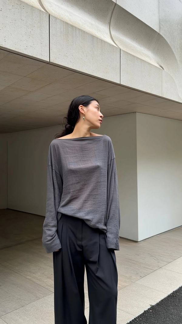 A casually candid iPhone snapshot of a woman standing thoughtfully under the overhang of a modernist stone building, her gaze directed sideways with an unposed, natural expression. She wears high-waisted black structured pants paired with an oversized long-sleeve washed grey linen top, its natural fabric textures and relaxed drape clearly visible, complemented by simple, delicate earrings. Soft natural daylight softly illuminates the scene, casting gentle ambient shadows on the stone's textured surface, emphasizing architectural lines and folds of fabric. The minimal composition centers on precise posture and the casual tilt of her head, with a slightly tilted framing capturing the clean lines of the building and the tactile contrasts between stone, fabric grain, and skin texture, intensifying intimate Japandi-influenced authenticity.