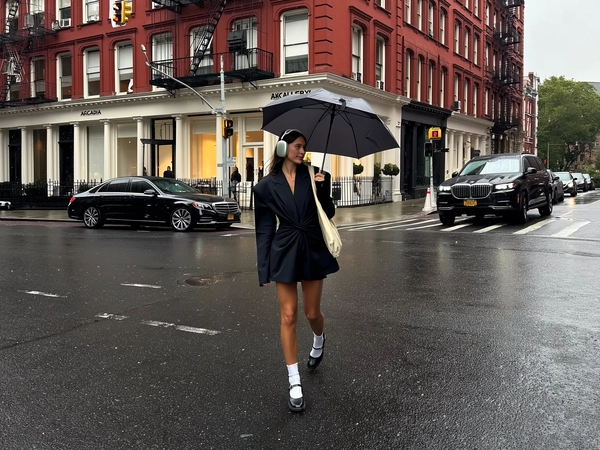 She crosses a slick city street under a soft drizzle, right hand gripping a dark grey umbrella while large white headphones cocoon her ears; an oversized black blazer cinched like a dress drapes over bare legs, paired with crisp white ankle socks and chunky black Mary Jane heels, a beige canvas tote swinging gently from one shoulder. Behind her, a deep red brick building with sharp white window trims and black iron fire escapes anchors the scene, storefront signs 'ARCADIA' and 'LKGALLERY' subtly visible above two parked black luxury sedans. The muted overcast light diffuses evenly, softening shadows across the wet asphalt that gleams with mirrored reflections of the quiet street. Tiny raindrops bead the umbrella fabric, slight motion blur brushes her mid-stride pose; the frame is portrait-orientated, eye-level and slightly off-center, lens catching the subdued city palette with natural iPhone sensor grain and the occasional raindrop distortion along the edges —rainy city snap, shot on iPhone