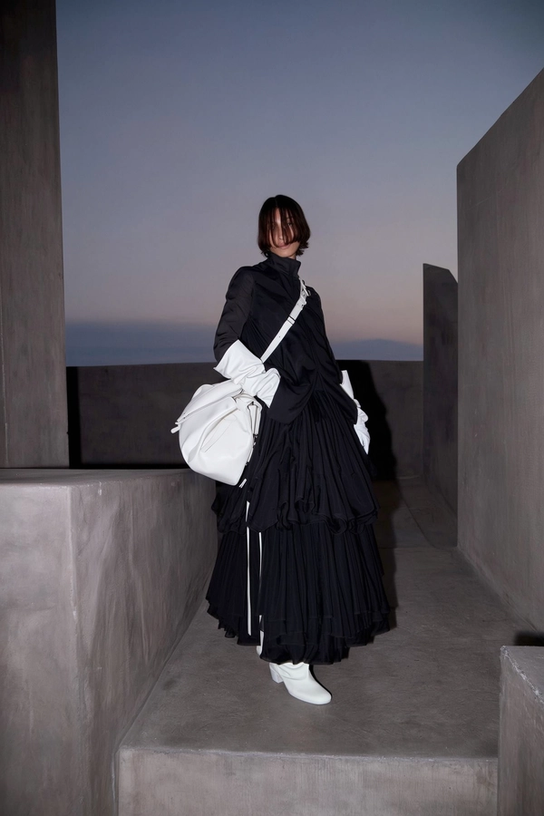 A softly dim-lit iPhone photograph capturing a model in an avant-garde Carol Christian Poell outfit, dressed in a flowing layered black skirt paired with sculptural white boots and oversized white leather bag and matching gloves. The figure stands slightly off-center within a Brutalist space defined by muted greys and shadowy charcoals, angular concrete formations, and subtle textural details. Twilight-inspired diffuse lighting casts gentle, soft shadows enhancing matte fabric folds and the quiet imperfections of the concrete. The composition is elevated and slightly tilted downward, providing generous negative space around the model and evoking intimate, introspective calmness amid the cool dusk hues.