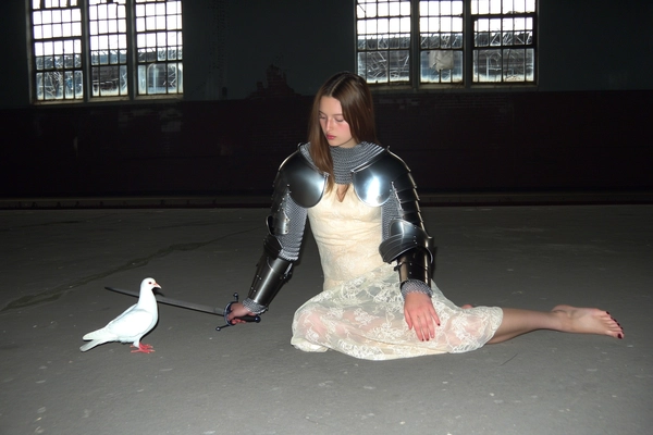 A wide, full-body, eye-level shot from an early 2000s-style digital camera explicitly capturing a young woman sitting gracefully on the floor of a long-abandoned train station with cracked windows. She wears minimal medieval armor consisting of shining shoulder guards and chainmail sleeves paired elegantly with a vintage lace dress, barefoot. Her calm, composed expression looks thoughtfully downward, sword resting elegantly by her side on the floor. A white dove stands quietly beside her, adding to the scene's contemplative mood. Dim, muted, natural light pours through the cracked windows mixing with harsh direct flash, emphasizing the textures of shiny metal armor, delicate lace fabric, bare skin, and individual hair strands. Slight digital noise and over-sharpening accentuate a gritty yet refined atmosphere. The full-body wide shot and informal but balanced framing capture the solemn medieval-core elegance, blending the dark, moody atmosphere of the abandoned interior with refined knightly poise characteristic of early 2000s digital photography.
