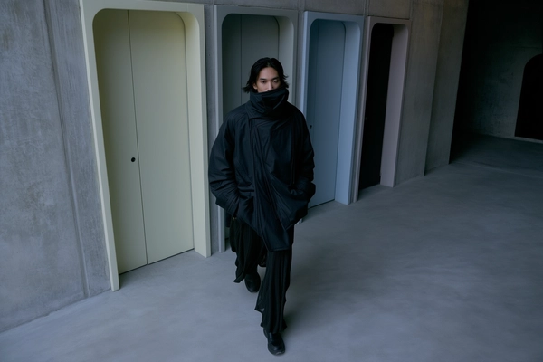 A softly dim-lit, dusk-inspired iPhone photograph capturing a man dressed in a layered avant-garde Julius outfit featuring a high-neck jacket partially covering the face and a long textured lower garment. He walks slowly down a Brutalist hallway softly illuminated by cool, muted dusk lighting, the concrete walls subtly textured in shadowy greys and charcoals. Freestanding door frames of varied, muted pastel colors provide gentle contrast, their minimal forms echoing quiet geometric lines. The model's hands rest hidden within the voluminous folds of the coat, his neutral expression and minimalist hairstyle contributing to an intimate, reflective mood. Shot from a slightly elevated, downward-tilted perspective, the composition places him casually off-center amid expansive negative space, softly framed by raw concrete surfaces textured with quiet imperfections, evoking relaxed asymmetry and refined textural realism.