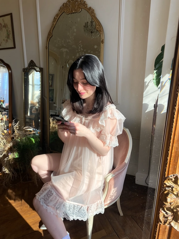 A softly radiant moment gently captured on an iPhone, presenting a pale-skinned young woman seated gracefully on a plush velvet chair within a sunlit room adorned with antique, ornate mirrors. She wears an airy, sheer lace babydoll dress characteristic of Jenny Fax, featuring delicate scalloped lace trims, subtle ruffles along the hem, and softly puffed sleeves. Her straight black hair is parted simply, framing her serene and introspective expression with minimal, fresh makeup enhancing her natural glow. Warm afternoon light filters through softly, illuminating the intricate lace texture, floating dust motes, and the gently aged surfaces of the antique mirrors. The composition is framed with a slight tilt and elevated angle, embracing a quietly intimate, whimsical vintage charm that harmonizes with Jenny Fax's dreamy, delicately feminine aesthetic.