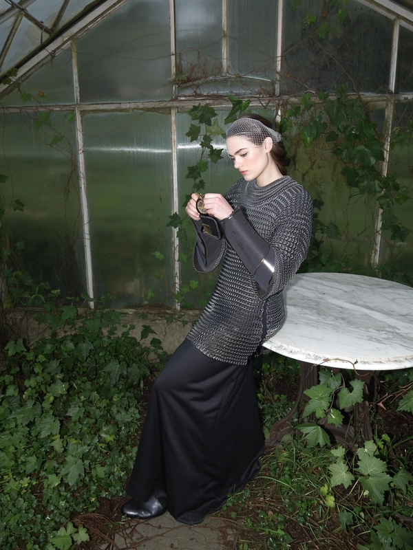 A wide, full-body, eye-level shot from an early 2000s-style digital camera capturing a young woman elegantly leaning gently against a cracked marble table inside a dim, fog-mutated greenhouse overtaken by vines. She wears dark chainmail armor blending with lush green foliage and a delicate mesh coif softly wrapping her head. Her composed, solemn gaze is directed downward, inspecting a broken pocket watch held thoughtfully in her hands. Muted morning fog presses against the greenhouse glass, creating a dim, diffused lighting enriched by the harsh direct flash that subtly picks out the shining textures and minor imperfections of her chainmail and fabric beneath. The framing is carefully balanced, emphasizing the melding of medieval refinement with nature's reclaiming ambiance, rendered in the gritty, high-contrast, desaturated palette characteristic of early 2000s digital photography fitting the medieval-core aesthetic.