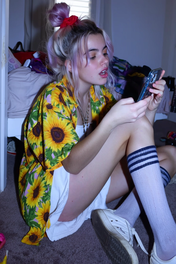 A candid mid-2010s style indoor snapshot of a young woman with subtle pastel pink and lilac ombré hair casually styled into a messy bun held by a red scrunchie. She sits on the floor in front of a slightly dirty mirror wearing a vibrant Hawaiian sunflower shirt layered over a white mini-dress, paired with long striped tube socks and worn white sneakers. Her lips are parted as she gazes into her smartphone with a relaxed, authentic expression. The dimly lit bedroom behind her reveals an unmade bed and scattered personal items, adding to the casual disarray. Muted ambient indoor lighting bathes the scene, featuring softly lowered contrast and cool-toned lavender-gray shadows. Visible textures include realistic skin imperfections, loosely defined pastel hair strands, detailed fabric grain of cotton shirt and socks, and subtle scuffs on the sneakers. The framing is informally off-center with a slight tilt, capturing an intimate, perfectly imperfect snapshot reminiscent of mid-2010s spontaneous youth photography aesthetics characterized by muted saturation, softened contrasts, and cool-toned shadow hues.