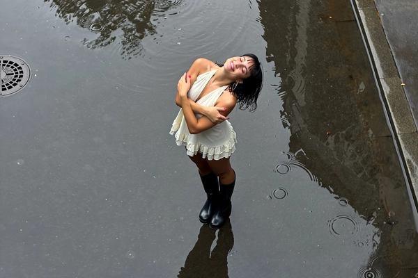 From above, she stands alone on glossy wet concrete, her off-white short halter dress layered with delicate ruffles contrasted by tall black leather boots grounded firmly amid puddled reflections. Arms folded gently over her chest, her head tilts back with eyes closed, a slight smile catching the overcast light filtering through misted air. Damp tendrils cling to her short dark hair, and large hoop earrings glint faintly. Around her, faint cracks and a street drain break the smooth dark surface, while diffuse daylight softens every ripple in the puddles. The quiet, cinematic moment feels suspended in time, the iPhone’s slight sensor grain and tiny motion blur lending raw intimacy to the scene. Framed from a high vantage point with a thoughtful Dutch tilt, the wet textures and her reflective embrace of rain compose a tender urban reverie—rainy city snap, shot on iPhone