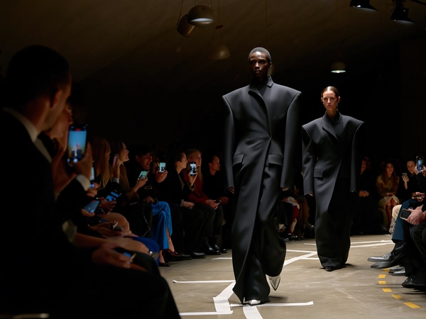 Two models stride down a slender, shadowed runway, each cloaked in jet-black tailored coats with dramatically exaggerated shoulders that sculpt their frames into powerful silhouettes. The dim venue narrows around them, with faint pools of amber light catching the sharp fabric creases and subtle sheen of fine wool. Their focused expressions cut through the low light as the second model follows close behind, heels clicking on the worn concrete floor marked with taped sight-lines. Overhead, sparse tungsten fixtures hang like dim sentinels, casting moody highlights that bloom softly on the coats’ structured seams. Guests flank the tight catwalk on narrow benches, phones lifted discreetly mid-whisper, their faces blurred in soft focus beyond the lead model’s poised stride. The iPhone’s sensor picks up mild digital noise in deep shadows, a slight hand-held tilt adding raw immediacy to the frame as fabric edges ripple with each step. Shot from an intimate front-row vantage point, the compressed space and interplay of form and darkness pulse with editorial tension—contemporary runway snapshot, shot on iPhone