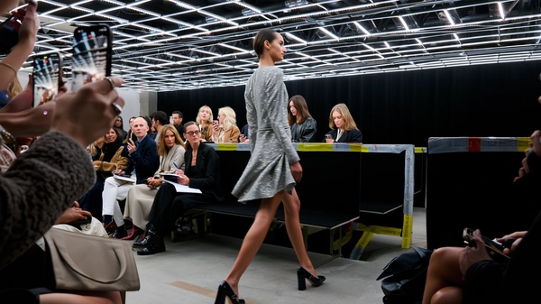 A sleek model glides past the front row clad in a shimmering silver mini-dress, its sequined surface catching fragmented light and throwing subtle sparkles onto polished leather pumps. Her confident stride kicks a hint of motion blur into the asymmetrical hemline, while her poised face reflects cool determination. Surrounding her, fashion editors in muted-toned cashmere and silk ensembles scribble swiftly in leather-bound notebooks, their concentrated expressions lit by softened tungsten spill from overhead fixtures. The front-row bench, upholstered in midnight velvet, faces a stripped-back industrial runway where scuffed tape markings and concrete grain add grit beneath footfalls. Above, grids of bright LEDs clash with the creeping shadows cast by delicate wire rigging, creating a contrast that the iPhone lens softens with gentle bloom and subtle sensor grain. The capture, taken from an angled eye-level seat, tilts slightly to include glimpses of nearby raised phones and murmuring guests, sealing the candid electric pulse of the moment—contemporary runway snapshot, shot on iPhone