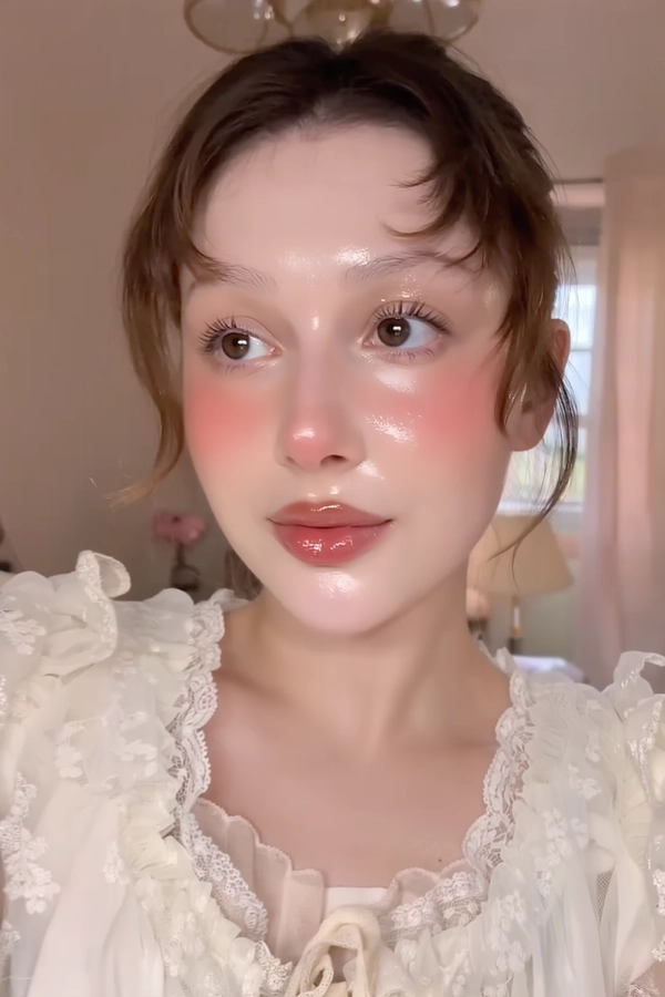 A close-up iPhone-style portrait of a young woman with soft red contouring gently blended across her cheeks and beneath her eyes, creating a subtle, artistic flush. Her skin is glossy and dewy with a natural, glass-like finish highlighting the nose, cheeks, and forehead, showing visible pores and authentic texture. Her eyes are expressive and glossy, enhanced with shimmery pastel eyeshadow in warm pink tones, and her natural-colored eyebrows are softly brushed upward. Her lips are slightly parted, coated with a high-gloss soft pink lip gloss that reflects the ambient light with moist, tactile shine. Her hair is natural and softly textured with loose strands framing her face, styled lightly with soft volume. She wears a delicate, vintage-inspired babydoll blouse in pale cream with puff sleeves, lace trims, and a subtle sheer overlay near the shoulders. The photo is tightly framed from just above her head to just below the collarbones, taken at a casual off-center angle with a slight tilt, conveying spontaneity. The background is softly blurred in pastel peach and cream hues, suggesting indoor ambient light mixed with gentle window daylight and faint phone flash, casting natural highlights and soft shadows. Fine details such as light flyaway hairs, skin texture, fabric stitching, and gloss shimmer particles are crisply visible, resulting in an intimate, authentic snapshot.