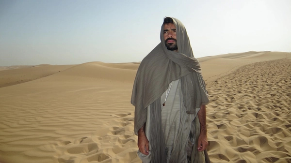 A candid, casually captured iPhone-style image of a Dutch man wearing layered, flowing earth-tone fabrics and a wide, draped scarf loosely wrapped around his neck and shoulders. He stands still in a subtly dramatic, upright pose within an expansive desert dune field characterized by soft, undulating sand dunes under natural sunlight. His face is partially shadowed by the scarf, creating a mysterious and introspective mood. The lighting is bright and natural with low contrast and subtle shadows, enhanced by the faint glow of an iPhone flash providing a spontaneous feel. The composition is minimalist and slightly asymmetrical, emphasizing the vastness of the desert and the tactile texture of the layered fabrics alongside the granular sand. Textural details of the rough dunes, soft drapery, and subtle skin nuances are captured with the authentic visual softness typical of casual iPhone photography.