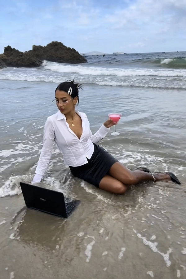 With waves lapping rhythmically against the shore, a stylish officewoman sits casually at the water's edge, her black pencil skirt soaked where it brushes the wet sand and shallow surf. She wears a crisp white blouse, unbuttoned just enough to suggest ease, and silver barrettes pin her wind-tossed hair neatly back. Her sharp gaze peers over the rims of slightly slid-down glasses at a black laptop precariously resting on damp beach sand, a pink cocktail in a martini glass balanced effortlessly in one hand. The bright midday sun glares off the rippling waves and jagged rocks framing the horizon, casting reflections that dance over the scene. Nearby, an old beige desktop monitor and full-sized keyboard lie half-submerged, underscoring the absurd twist on remote work culture. The photo has a grainy quality, suggesting it may be a still from a video or taken with an older digital camera. Quick snapshots create a soft-focus effect at the edges, and muted colors swirl into a nostalgic haze, capturing the unlikely marriage of corporate formality and beach tranquility. A gentle breeze ruffles her wet skirt as the salt spray mingles with office paraphernalia—early-2000s digicam snapshot, grainy quality.