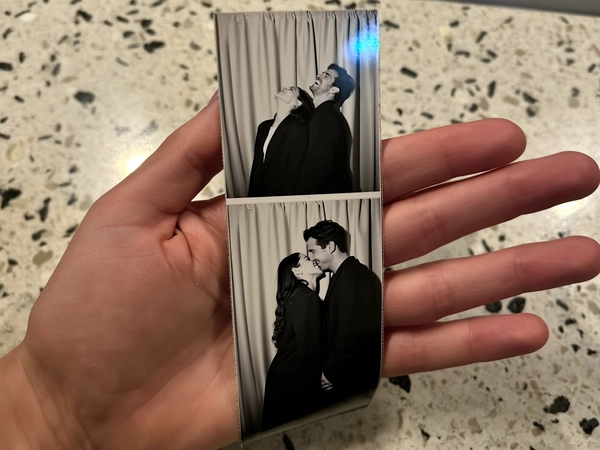 A narrow vertical photostrip lies gently cupped in an open palm above a speckled terrazzo countertop, the hand relaxed and casual. The black-and-white images reveal a couple clad in long coats, each frame capturing a shift in mood: the first upright and composed, the woman tilting her head back in soft relief, the man bursting into laughter, finally their foreheads touching in a tender, quiet embrace. Behind them, a neutral beige curtain with subtle folds frames the scene with photobooth authenticity. The strip shows faint creases and gentle fingerprints, adding tactile realism. Soft shadows and a delicate iPhone flash reflection create muted contrast with grainy sensor noise, while the composition rests slightly skewed, as if casually snapped mid-moment, evoking a nostalgic, playful intimacy—contemporary photobooth snapshot, shot on iPhone.