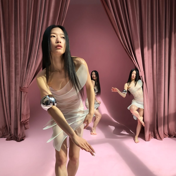 A spontaneous and surreal iPhone photograph of a woman duplicated multiple times in the exact same translucent sculptural dress paired with shiny metallic cuffs, set against a soft pink theater backdrop with rich, heavy velvet curtains. Each figure engages in a subtly different pose: one reaches forward with a delicate hand, another gracefully balances on one leg, while a smaller-scale version peeks curiously from behind the curtain. Natural diffused daylight gently casts realistic shadows and soft highlights on the fabric's translucent textures and metallic accessories, emphasizing fine skin details and hair strands. The framing combines close-up and distant perspectives with intuitive casual cropping, evoking a whimsical yet authentic candid moment where surreal repetition and theatrical environment intertwine seamlessly.