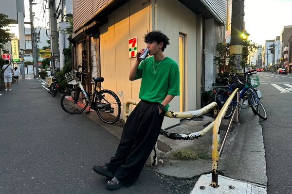 A casually stylish young man drifts in early evening hues, loose green tee blending softly with oversized black trousers as he perches on a chipped sidewalk railing outside a weathered narrow building. A silver chain glimmers faintly under gentle ambient glow while he sips canned coffee mid-scroll, unposed and absorbed. Behind, a familiar 7-Eleven neon sign hums low, bikes clustered in quiet linear disarray. The iPhone’s natural warmth tames city colors without softening the cracked asphalt beneath scuffed shoes. Shadows balance perfectly against fading daylight, creasing fabric folds and tousled hair with honesty. The frame tilts slightly off-center, lens flare whispering from distant LEDs, grounding him in this honest Tokyo moment where streetwear meets everyday rhythm—Tokyo street snap, shot on iPhone