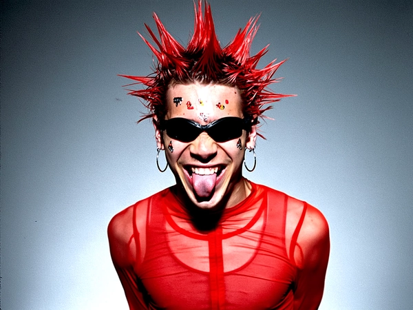 Medium close-up shot of a guy with bright cherry-red spiked hair styled into sharp points, scattered with small face stickers across his cheeks and forehead. He wears slim, black wraparound goggles pushed slightly over his eyes, lending an underground cyber-rave vibe. His sheer, neon orange athletic top clings tight and reveals subtle muscle definition beneath. Large silver hoop earrings dangle from both ears, catching the ambient studio lights. His mouth is open mid-laugh with his tongue out, flashing a manic grin that channels the wild confidence of MySpace-era youth culture. The backdrop is a cold grey with a gritty, grainy texture that frames him starkly without distraction. Posed deliberately with a defiant lean forward, eyes half-shielded behind the goggles, the image radiates electric attitude and controlled chaos under harsh artificial lighting. —casual candid early-2000s Y2K snapshot, grainy softness