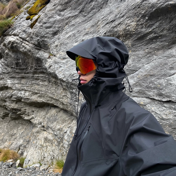 A naturally posed side profile of an individual standing relaxed in front of a textured rugged rock wall within a mountainous terrain. The person wears a matte black technical waterproof hardshell jacket with an oversized hood fully raised, featuring angular shaping and cinch cords creating a sculptural, structured form that closely envelops the head. The jacket shows taped seams and waterproof zippers, highlighting its high-performance, utilitarian design. The individual sports high-contrast, orange-tinted wraparound goggles that vividly reflect the surrounding natural alpine landscape, partially obscuring the face so only the nose and part of the lips are visible beneath the hood and eyewear. Soft, diffused, indirect daylight gently illuminates the scene, emphasizing the rich textures of the waterproof fabric and the coarse rocky surfaces behind. The casual, spontaneous framing includes a subtle, slightly tilted angle, reinforcing the authentic candid iPhone photography aesthetic. This hyper-realistic, cinematic image conveys a calm, tactical, futuristic outdoor vibe within a rugged mountain setting, seamlessly blending minimalist mountain utility with cyberhiker fashion.