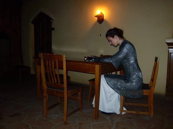 A wide, full-body, eye-level shot from an early 2000s-style digital camera explicitly capturing a woman seated gracefully at a simple wooden table inside a dimly lit monastic room. She wears dark chainmail combined elegantly with contemporary clothing, with no gauntlets, her posture composed and solemn, head slightly bowed in contemplation, hands resting gently on the table near a small cup. The only light source is a warm wall sconce casting muted, dim illumination, creating a reverent atmosphere with heavy shadows and rich textures. The harsh direct flash characteristic of early 2000s digital photography subtly enhances the sheen of chainmail and the natural skin and fabric textures, adding visible digital noise and slight over-sharpening. The composition is formally balanced and symmetrical, the full-body wide shot emphasizing the serene knightly mood. The overall image blends medieval-core fashion influences with a dark, moody, and gritty but elegantly restrained visual tone typical of early 2000s digital captures.